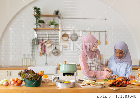 Joyful cooking session lifestyle in contemporary kitchen, Muslim women and girl of different ages in hijabs chop fresh vegetables and prepare meal amidst colorful ingredients and kitchenware. 131283049