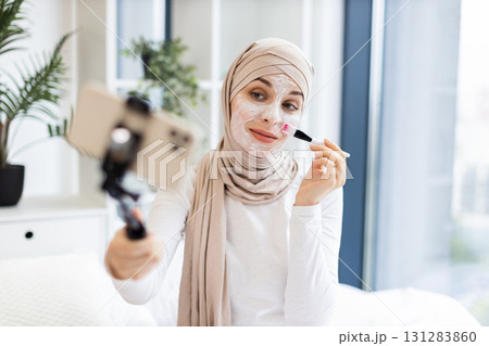 Young Muslim woman recording skincare vlog applying facial mask using brush in casual indoor setting. Promoting self-care, beauty, and skin health concepts with relaxed and motivating atmosphere. 131283860