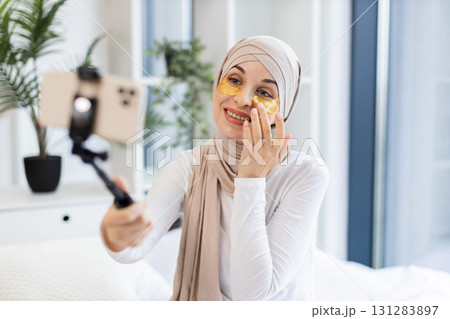 Muslim woman wearing hijab applying gold hydrogel patches under eyes recording vlog indoors Muslim woman wearing hijab applying gold hydrogel patches under eyes recording vlog indoors 131283897