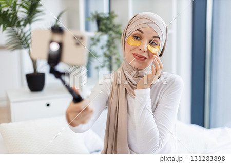 Muslim woman wearing hijab applying gold hydrogel patches under eyes recording vlog indoors Muslim woman wearing hijab applying gold hydrogel patches under eyes recording vlog indoors 131283898