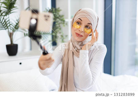 Young Muslim woman wearing hijab records vlog with smartphone while using gold eye patches for skincare in modern home setting. Concept of beauty, self-care, and digital content creation. 131283899