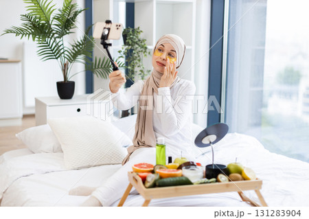Young Muslim woman wearing hijab recording vlog about using gold under-eye patches. She sits in a bright bedroom with skincare products and fruits on display, sharing beauty tips with her audience. Young Muslim woman wearing hijab recording vlog about using gold under-eye patches. She sits in a bright bedroom with skincare products and fruits on display, sharing beauty tips with her audience. 131283904