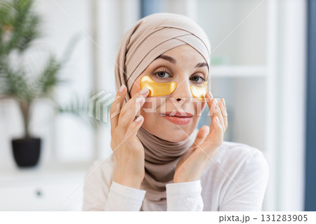 Young Muslim woman wearing headscarf applying gold hydrogel patches under her eyes, emphasizing skincare and self-care practices, photographed in modern environment promoting beauty, health Young Muslim woman wearing headscarf applying gold hydrogel patches under her eyes, emphasizing skincare and self-care practices, photographed in modern environment promoting beauty, health 131283905