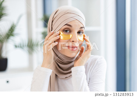 Young Muslim woman in beige hijab applying gold hydrogel eye patches for skincare in modern indoor environment. Promoting self-care and healthy lifestyle concept, model demonstrates beauty. Young Muslim woman in beige hijab applying gold hydrogel eye patches for skincare in modern indoor environment. Promoting self-care and healthy lifestyle concept, model demonstrates beauty. 131283913