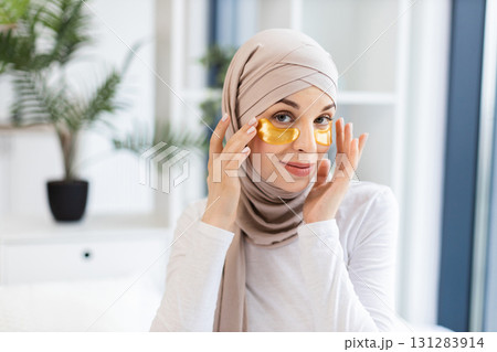 Young Muslim woman in beige hijab applying gold hydrogel eye patches for skincare in modern indoor environment. Promoting self-care and healthy lifestyle concept, model demonstrates beauty. Young Muslim woman in beige hijab applying gold hydrogel eye patches for skincare in modern indoor environment. Promoting self-care and healthy lifestyle concept, model demonstrates beauty. 131283914