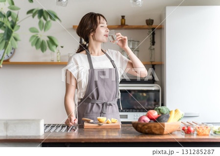 キッチンでコップで水を飲むエプロン姿の女性（水分補給,ミネラルウォーター,ウォーターサーバー） 131285287