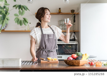 キッチンでコップで水を飲むエプロン姿の女性（水分補給,ミネラルウォーター,ウォーターサーバー） 131285288