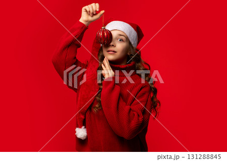 Girl in Santa hat holding red Christmas ornament near eye. Girl in Santa hat holding red Christmas ornament near eye. 131288845