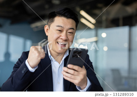 Close-up photo of a young Asian man in a business suit sitting at a desk in the office, looking at the phone screen and showing a victory yes gesture with his hand. Close-up photo of a young Asian man in a business suit sitting at a desk in the office, looking at the phone screen and showing a victory yes gesture with his hand. 131289310