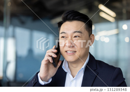 Close-up photo of a young Asian businessman standing in an office center and talking on a mobile phone. Close-up photo of a young Asian businessman standing in an office center and talking on a mobile phone. 131289348