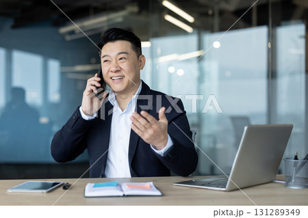 Smiling young Asian businessman in suit sitting in office at desk and talking while gesturing with hands. Smiling young Asian businessman in suit sitting in office at desk and talking while gesturing with hands. 131289349