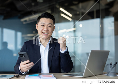 Portrait of a smiling young successful businessman sitting at a desk in the office, holding a mobile phone and showing a victory gesture with his hand. Portrait of a smiling young successful businessman sitting at a desk in the office, holding a mobile phone and showing a victory gesture with his hand. 131289360