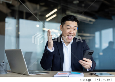 A happy Asian man in a business suit is sitting in the office at the table and is happy looking at the screen of the mobile phone he is holding in his hand. 131289361
