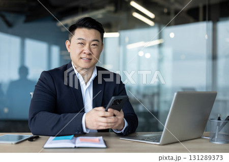 Portrait of a young smiling Asian businessman sitting at his desk in the office, holding a mobile phone and looking at the camera. 131289373