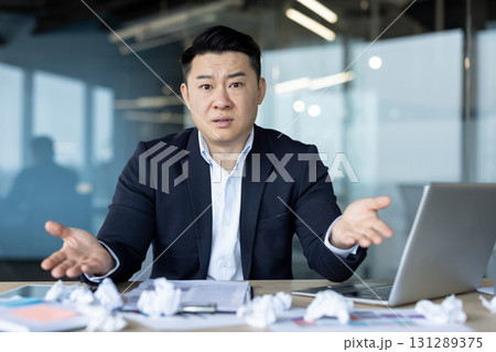 Portrait of a young Asian man in a suit sitting in an office at a desk with crumpled papers scattered on it, folding his hands in frustration and looking at the camera. Portrait of a young Asian man in a suit sitting in an office at a desk with crumpled papers scattered on it, folding his hands in frustration and looking at the camera. 131289375