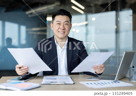 Portrait of a young Asian businessman working in the office with a laptop and documents, sitting at a desk, holding papers and looking at the camera. 131289405