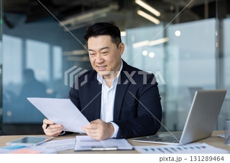 Asian young smiling male businessman sits in the office at a desk with a laptop in a business suit and reads documents in his hands. 131289464