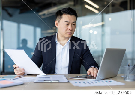 A serious young Asian businessman is sitting in the office at a desk and is concentrating on working on a laptop, holding documents and typing on a keyboard. 131289509