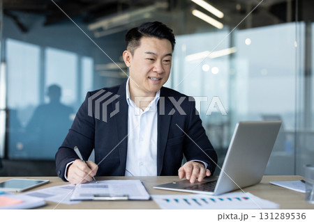 Smiling young Asian man working in the office with documents and a laptop. 131289536
