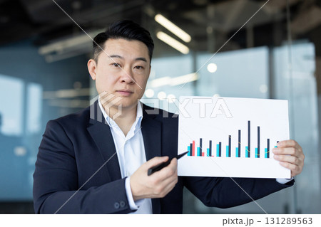 A serious young Asian man in a business suit is working in the office, sitting at the workplace and pointing at the camera with a pen to the graphs and documents in his hands. 131289563