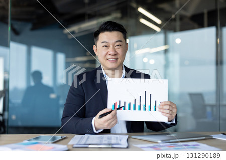 Portrait of a young smiling Asian man sitting in the office at a desk and showing documents with company graphs and revenues to the camera. Portrait of a young smiling Asian man sitting in the office at a desk and showing documents with company graphs and revenues to the camera. 131290189
