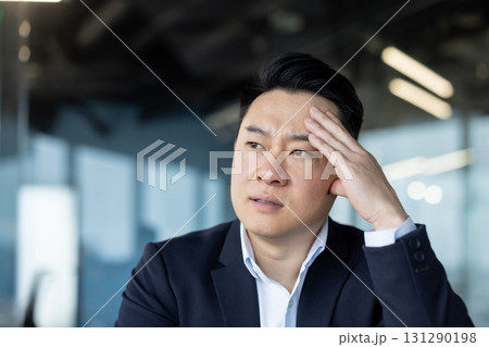 Close-up photo of a young Asian man in a suit sitting at a desk in the office and holding his head, feeling pain, thinking about solving a problem. Close-up photo of a young Asian man in a suit sitting at a desk in the office and holding his head, feeling pain, thinking about solving a problem. 131290198