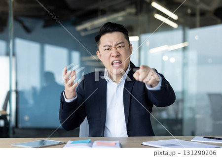 Portrait of angry young Asian man in business suit sitting at desk in office, talking and pointing aggressively at camera. Portrait of angry young Asian man in business suit sitting at desk in office, talking and pointing aggressively at camera. 131290206