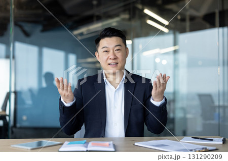A professional Asian man in a dark suit gestures while addressing an audience, possibly a meeting. 131290209