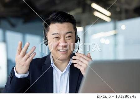 Close-up photo of a smiling young Asian man in a suit and headset sitting in the office ,at a laptop and talking on a video call. Close-up photo of a smiling young Asian man in a suit and headset sitting in the office ,at a laptop and talking on a video call. 131290248