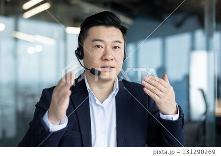 Close-up portrait of a young Asian man in a suit and headset talking to the camera and gesturing with his hands. Close-up portrait of a young Asian man in a suit and headset talking to the camera and gesturing with his hands. 131290269