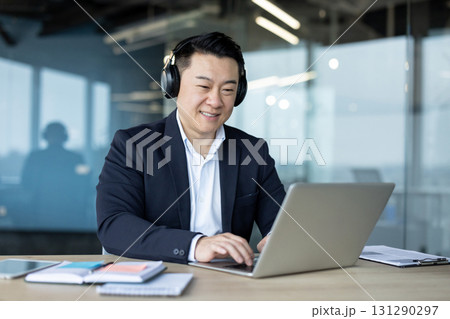 Asian young male businessman sitting in the office wearing headphones and smiling while working on a laptop. 131290297