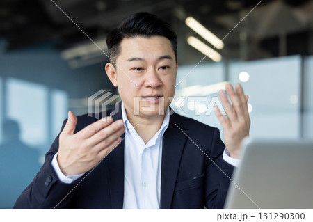 Asian young male businessman sitting in the office at the table and talking on a video call on a laptop, gesturing with his hands. Asian young male businessman sitting in the office at the table and talking on a video call on a laptop, gesturing with his hands. 131290300