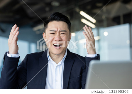 An upset young Asian man sits in the office at a desk in front of a laptop, looking at the screen in frustration, spreading his arms. An upset young Asian man sits in the office at a desk in front of a laptop, looking at the screen in frustration, spreading his arms. 131290305