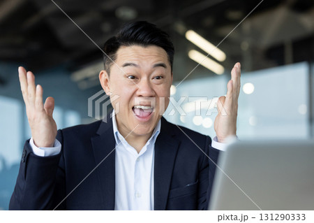 An excited asian businessman celebrates a victory or good news while working. He is looking at his laptop. An excited asian businessman celebrates a victory or good news while working. He is looking at his laptop. 131290333