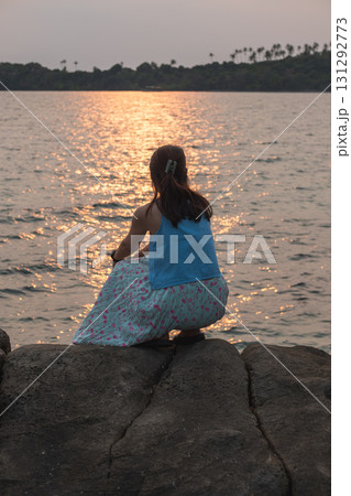 Asian woman sitting on rocks watching sunset over calm sea with distant island 131292773