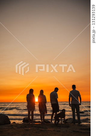 Four people standing with a German Boxer dog on the beach during a stunning sunset, looking at the ocean waves and enjoying a calm evening together. 131293139