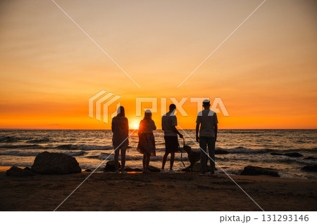 Group of four people with a German Boxer dog standing on the sandy beach at sunset, gazing at the ocean horizon during a peaceful evening walk. 131293146