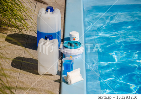 Swimming pool maintenance set with chemical canisters, chlorine tablets, floating dispenser, measuring cup with blue liquid and cleaning supplies placed on pool edge 131293210