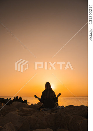 Silhouette of a young woman meditating by the sea at sunrise, sitting cross-legged on rocks, hands in mudra, symbolizing harmony, self care, emotional balance, and connection with nature 131293214