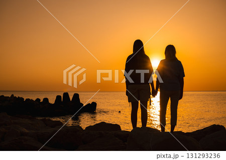 Two women holding hands on the rocks at sunrise, looking at the sea horizon, symbolizing love, unity, support, family bonds, and emotional well-being Two women holding hands on the rocks at sunrise, looking at the sea horizon, symbolizing love, unity, support, family bonds, and emotional well-being 131293236