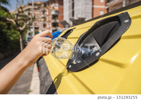 Close up of a hand placing a plastic bottle into a yellow recycling bin outdoors under bright sunlight. Close up of a hand placing a plastic bottle into a yellow recycling bin outdoors under bright sunlight. 131293255