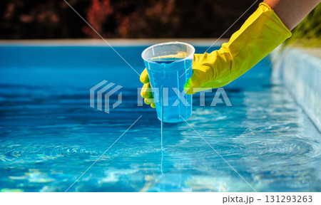 Hand in yellow protective glove pouring blue pool chemical from measuring cup into clear water for swimming pool disinfection and maintenance Hand in yellow protective glove pouring blue pool chemical from measuring cup into clear water for swimming pool disinfection and maintenance 131293263