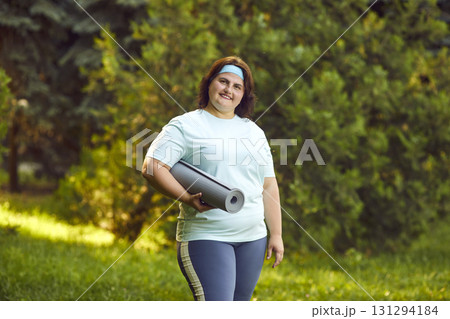 Fat happy woman going to do sport exercises holding yoga mat standing in summer park outdoors. 131294184