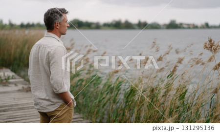 A contemplative man in a linen shirt stands on a peaceful pier gazing at the lake, surrounded by swaying grasses and moody skies, captured in warm vintage film style. 131295136