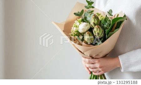 Close-up of hands holding rustic bouquet of wildflowers and artichokes in kraft paper, with soft cotton sleeves and neutral backdrop, captured in gentle window light for a natural, organic look. 131295188