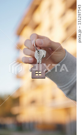 Vertical close up of male hand holding house key with home shaped keychain on background of modern apartment building. Symbol of property ownership, mortgage, new home and real estate deal. 131297543
