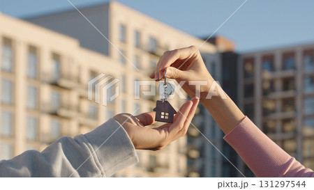 Close up of female real estate owner handing over house key with home shaped keychain to young man outdoors on background of modern apartment building. New home, first time buyer, and property deal. 131297544