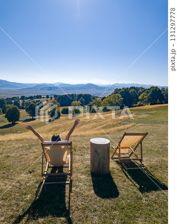 Side view of a woman sitting on a lounge chair with raised arms, overlooking hills and distant mountains in Durmitor 131297778