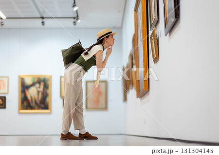 Wide shot of a young Caucasian woman in a straw hat and elegant clothes carefully contemplating modern art in the gallery. Low angle side view 131304145