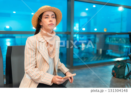 Mid shot of young Caucasian elegant woman is sitting in an airport lounge chair. Windows in background. Concept of boarding at terminal before flight 131304148